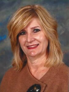 Amy Perry A woman with shoulder-length blonde hair and light skin smiles at the camera. She is wearing a brown top and has a neutral, blue-gray background behind her.