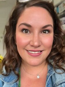 Beth Kirby A woman with wavy brown hair smiles at the camera. She is wearing a light makeup, a denim jacket, and a silver necklace. The background appears to be indoors with shelves and various items visible.