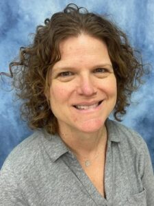 Christina-Tucker A person with short, curly brown hair smiles at the camera. They are wearing a gray collared shirt and a small necklace, with a blue and white mottled background behind them.