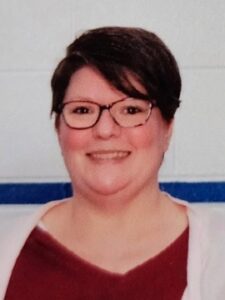Elizabeth Cox A person with short dark hair and glasses is smiling at the camera. They are wearing a maroon top and a light-colored cardigan, standing in front of a white wall with a blue horizontal stripe.