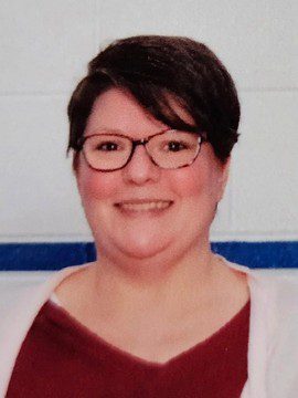 A person with short dark hair and glasses is smiling at the camera. They are wearing a maroon top and a light-colored cardigan, standing in front of a white wall with a blue horizontal stripe.