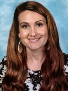 Jenna Milam A woman with long brown hair, wearing oval earrings and a floral-patterned top, smiles in front of a blue, cloudy background.