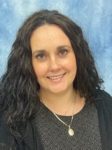 Jennifer-Camarillo-scaled A woman with long, curly dark hair smiles at the camera. She is wearing a patterned black top, a black cardigan, and a gold necklace. The background is a blue and white mottled backdrop.