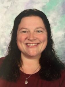 Julie Basham A woman with long dark hair, wearing a maroon top and a silver necklace, smiles in front of a gray and light green mottled background.