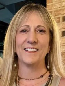Kristin Dixon A smiling woman with straight blonde hair, wearing dangling earrings and layered necklaces, stands in front of a brick wall.