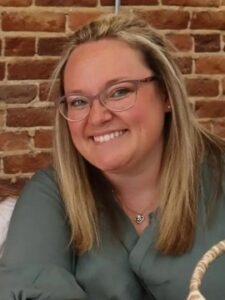 Screenshot A woman with straight blonde hair and glasses smiles at the camera. She is wearing a green top and a heart-shaped necklace, sitting in front of a brick wall.