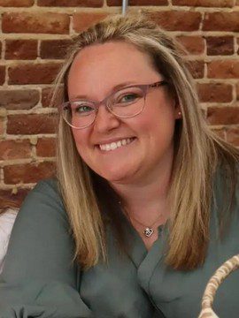 A woman with straight blonde hair and glasses smiles at the camera. She is wearing a green top and a heart-shaped necklace, sitting in front of a brick wall.