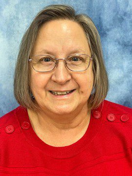 A smiling older woman with straight, shoulder-length gray hair and glasses, wearing a bright red sweater with buttons on the shoulders, in front of a blue marbled background.