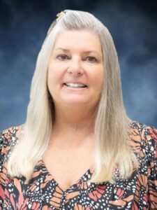 Mary Grace DePolo-Peck A smiling woman with long, straight, gray-blonde hair wears a patterned blouse and stands in front of a blurred dark blue background.