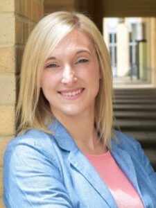 Megan Hatfield A woman with straight blonde hair, wearing a light blue blazer over a pink top, smiles while leaning against a brick wall in a sunlit corridor with columns in the background.
