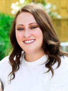 Sarah Worley A woman with long brown wavy hair, wearing a white top and a nose ring, smiling outdoors in front of greenery and a wooden fence.