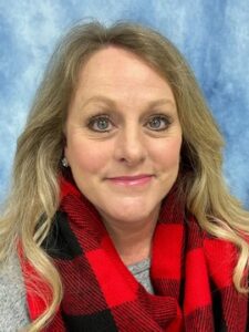 Stacy-Underwood-scaled A woman with long blonde hair smiles at the camera. She is wearing a gray top and a red and black plaid scarf. The background is a blue and white marbled pattern.