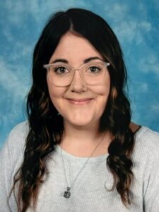 Screenshot A woman with long, wavy dark hair and glasses is smiling at the camera. She is wearing a light gray top and a necklace, with a blue mottled background behind her.