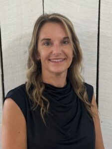 Traci Stacy A woman with wavy, light brown hair wearing a black sleeveless top smiles at the camera in front of a white wooden wall.