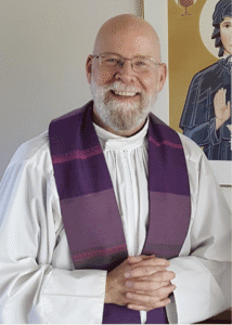 Screenshot 2026-07-14 at 11.17.14 PM A smiling man with a white beard and glasses wearing a white robe and a purple stole, standing in front of religious artwork, hands clasped together.