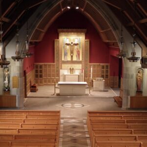 Interior of a church with wooden pews, a central altar, religious statues, and a large crucifix above the altar, set against a red background. High arched ceiling and symmetrical lighting fixtures are visible.