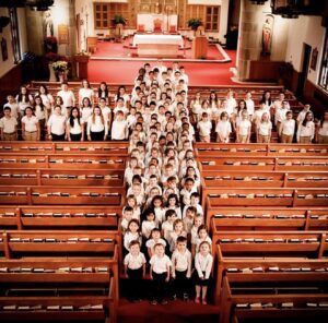 A large group of children and adults stand in church pews, arranged in the shape of a cross, all wearing white tops and dark pants, facing the camera in a well-lit sanctuary with a red carpeted altar.