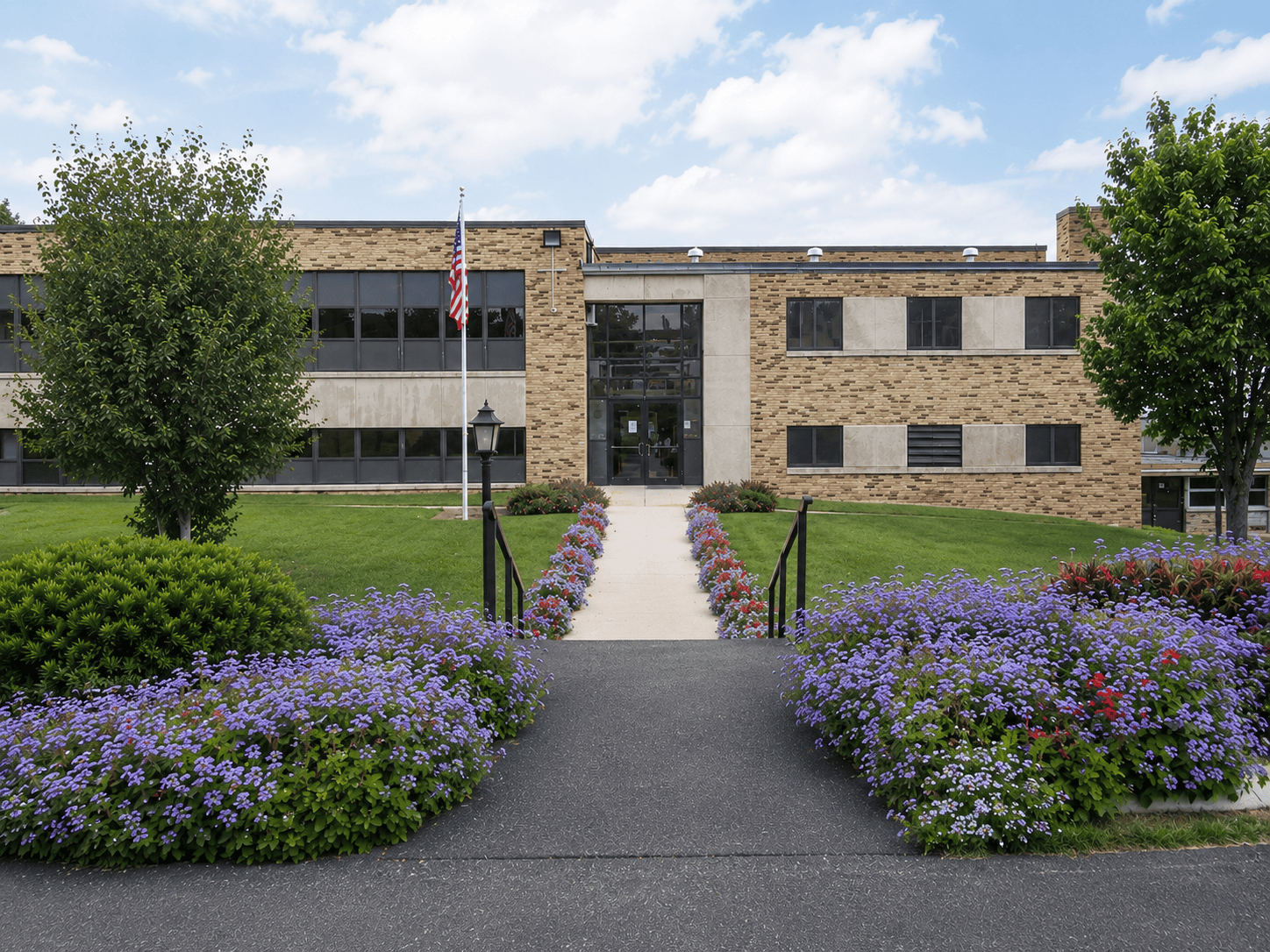 A two-story brick building with an American flag, surrounded by green lawns, trees, and vibrant flower beds lining a walkway leading to the entrance, under a partly cloudy sky.